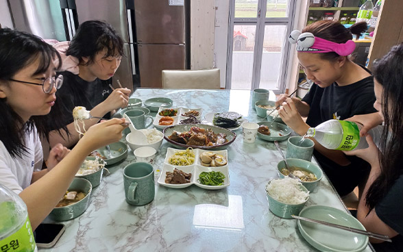 식탁 위 나물로 보이는 여러가지 반찬과 학생 4명이 마주 앉아 밥을 먹고 있는 모습